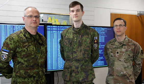 Three soldiers in camouflage outfits stand in front of two big screens.