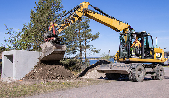 Keltainen kaivinkone kasaa maata vasemmalla olevan betonirakenteen seinustalle. Taustalla on kesäinen merimaisema.