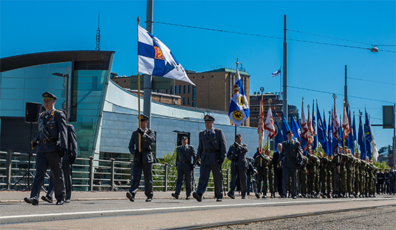 Puolustusvoimien kaikki puolustushaarat marsimmassa paraatissa. Etualalla kannetaan Suomen lippua. Taaemapana Puolustusvoimien eri toimijoiden lippuja.