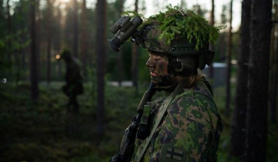 Etualalla suomalainen maastopukuinen sotilas, jonka kypärässä Senop-valonvahvistin. Etualan sotilas on kuvattu puolikuvana. Kuvan taustalla on toinen sotilas sekä hämärä kesäinen metsä.