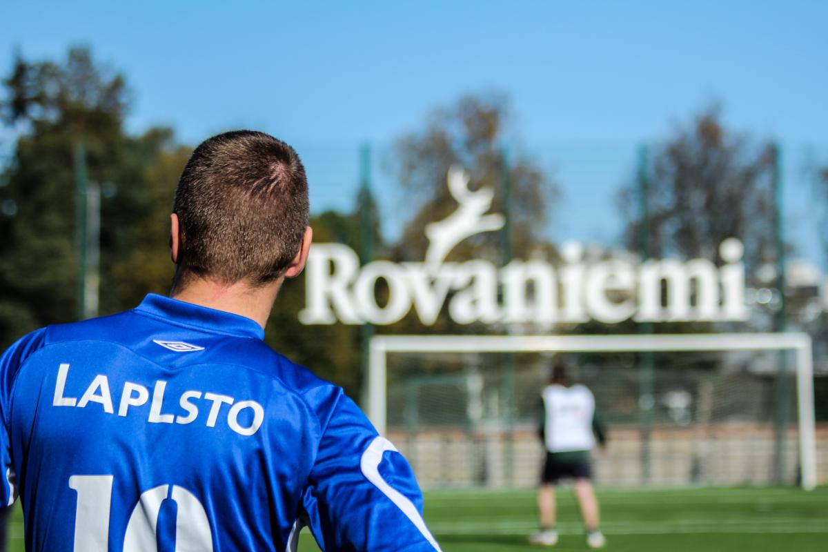 Mies seisoo selin kameraan, sininen jalkapallopaita päällä, selässä lukee LAPLSTO. Taustalla Rovaniemi-kyltti ja jalkapallomaali.