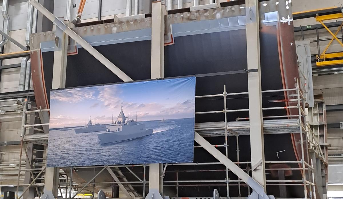 Kuvassa on monitoimikorvetin teräksinen köliosa, jonka edessä on isona julisteen havainnekuva valmiista aluksesta.