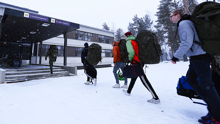 Siviiliasuisia nuoria kantamassa varustesäkkejä sisään kasarmirakennukseen. Maassa on lunta.