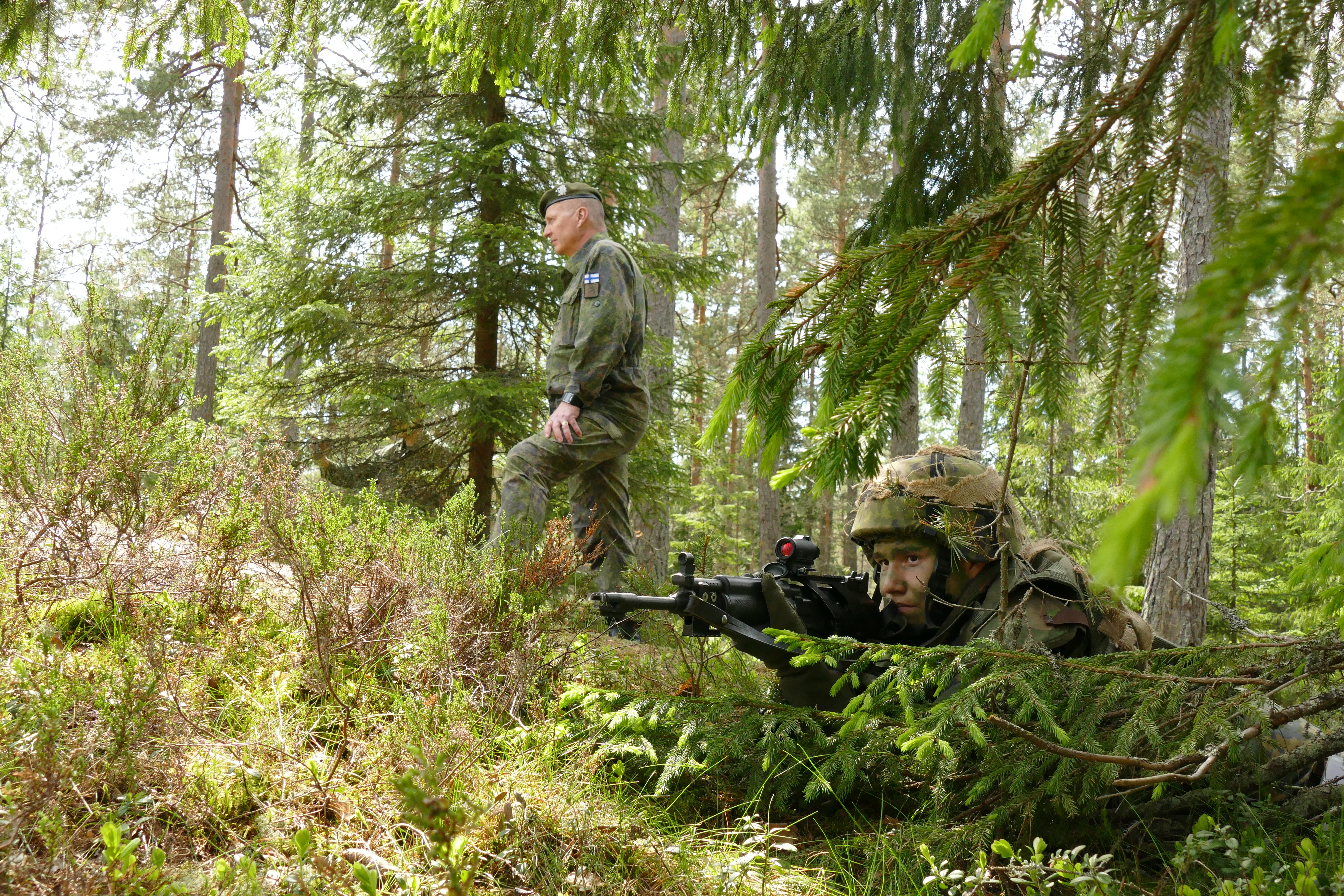 Etualalla RU-kurssilainen tähtää aseella, taustalla Maavoimien komentaja Hulkko tarkastaa Kärkikomppanian maastoharjoituksen.