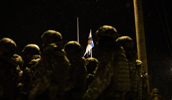 Etualalla pieni joukko varusmiehiä seisoo asennossa marraskuun pimeydessä. Taustalla heiluu lipputangossa Suomen lippu.