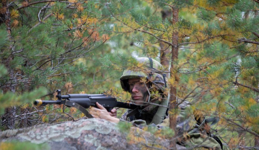 Sotilas kiven takana puiden suojassa, kasvot maalattuina, ase tuettuna kiveen.