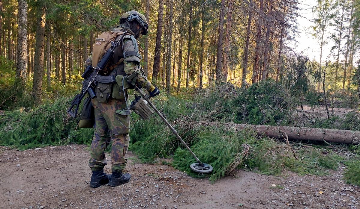 Sotilas metsätiellä, miinaharava käsissään haravoi maata tielle kaadettujen puiden luota
