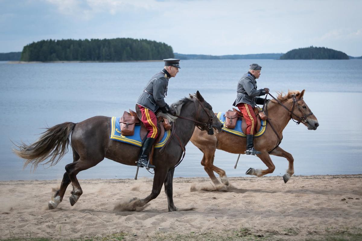 Kaksi ratsukkoa laukkaa järven rannalla perinnevarusteissa.