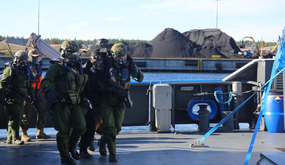 Kaksi sotilasta happimaskeissa auttaa haavoittunutta pois laivasta. Taustalla kaksi muuta sotilasta ja sataman kenttiä.