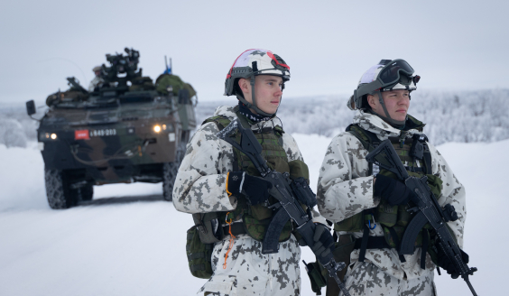 Kaksi varusmiestä seisoo taisteluvarustuksessa lumisessa maastossa. Taustalla kuljetuspanssariajoneuvo Patria XA-360.