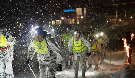 Lumipukuisia ja huomioliivisiä hiihtäjiä lumisateessa hiihtokilpailun yhteislähdön jälkeen hiihtämässä jätkänkynttilöiden palaessa taustalla.