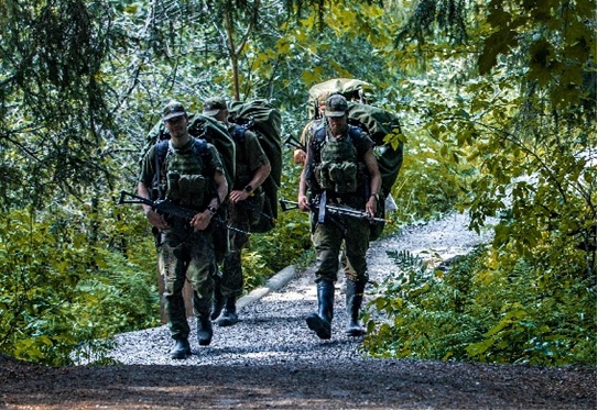 Urheilukoulun varusmiehet marssivat kesällä metsässä reput selässä ja aseet käsissä.