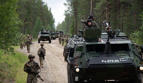 Viisi panssaroitua ajoneuvoa etenee metsän keskellä olevalla hiekkatiellä. Ajoneuvojen vierellä kulkee useita aseita kantavia sotilaita jalan.