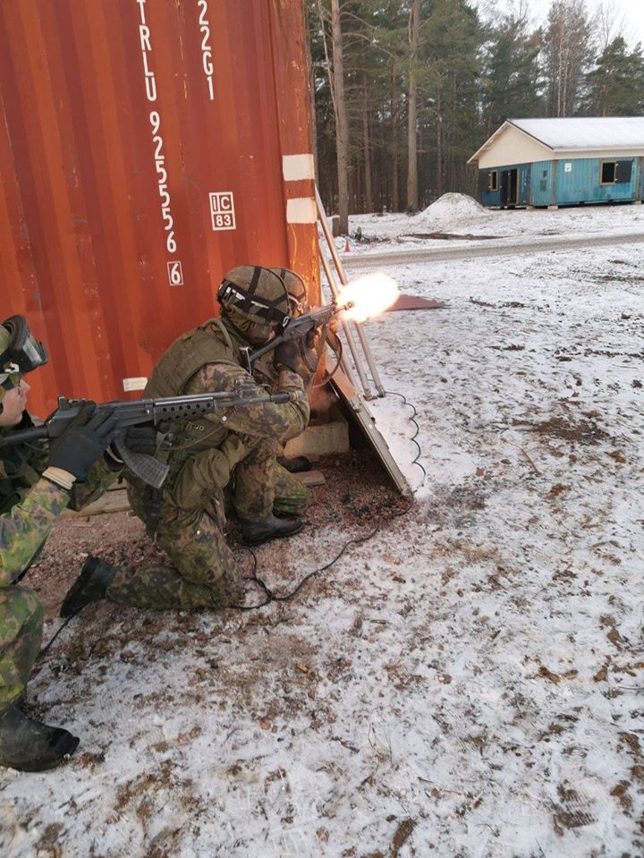 Jalkaväen sotureita ampumassa rynnäkkökiväärillä kulman takaa kohti sinistä rakennusta.