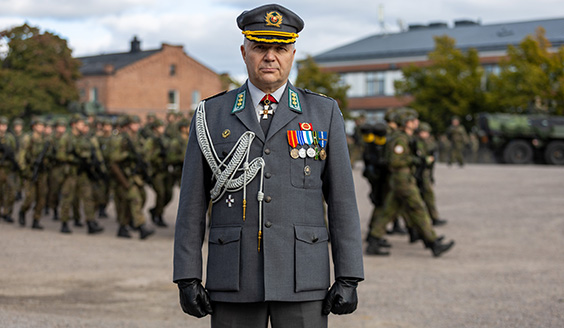 Eversti Ari Lehmuslehti Santahaminan paraatikentällä. Taustalla marssivia joukkoja ja kasarmirakennuksia.