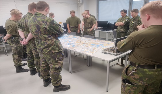 Yksitoista maastopukuissta reserviläistä seisoo pöydän ympärillä. Pöydällä on kartta ja pelimerkkejä.