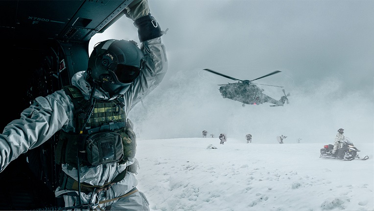 NH-90-helikopteri lähietäisyydellä ilmassa, takana näkyy moottorikelkkaileva sotilas sekä hiihtäviä sotilaita