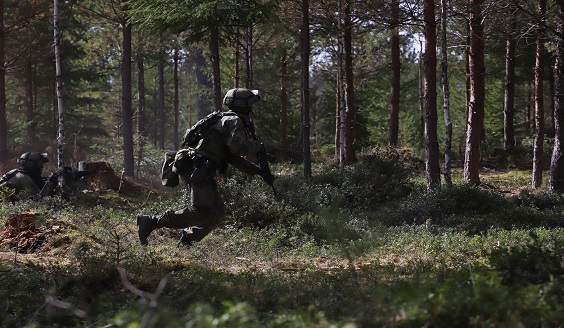 maastopukuinen varusmies juoksee vihreässä metsässä kantaen rynnäkkökivääriä