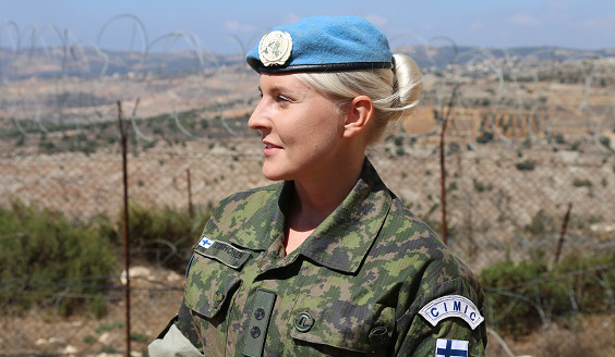 Luutnantti Jenny Korhonen. Taustalla korkea piikkilanka-aita ja hiekkainen maisema.