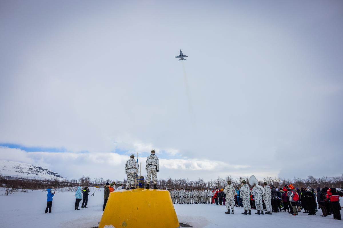 Hornet-hävittäjä lentää kolmen valtakunnan rajapyykin yli. Rajapyykillä runsaasti yleisöä ja sotilaita. Suomen lippu liehuu rajapyykillä.