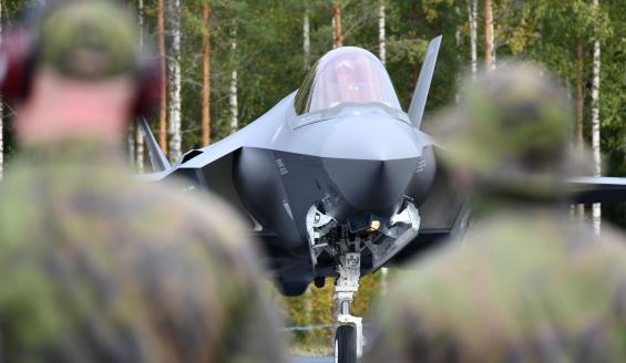 Kahden Suomen puolustusvoimien maastopukuun pukeutuneen henkilön takaa otettu kuva, jossa henkilöiden välissä näkymä tarkentuu norjalaiseen F-35A-hävittäjään, joka on juuri laskeutunut Tervon maantietukikohtaan ja rullannut levikkeelle tankattavaksi. Koneen keula osoittaa lähes suoraan katsojaa kohti.