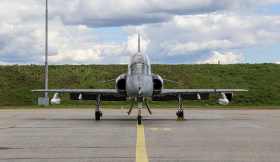 Suoraan koneen edestäpäin otettu kuva harmaasta Ilmavoimien Hawk-suihkuharjoituskoneesta. Kone on paikoillaan lentokenttäalueella taustallaan kirkaan vihreän ruohoinen maavalli ja pilvinen kesätaivas.