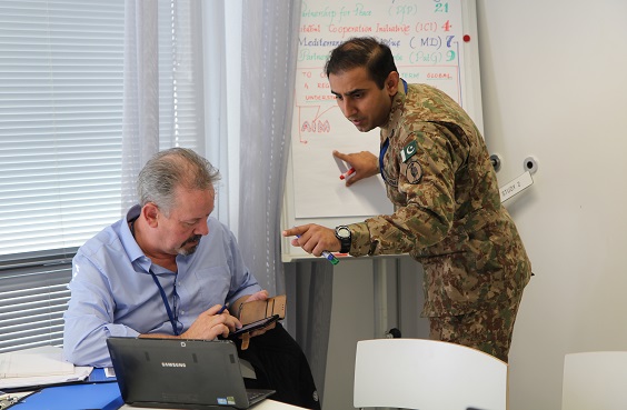 A foreign soldier is pointing at the whiteboard and a civilian is watching his phone
