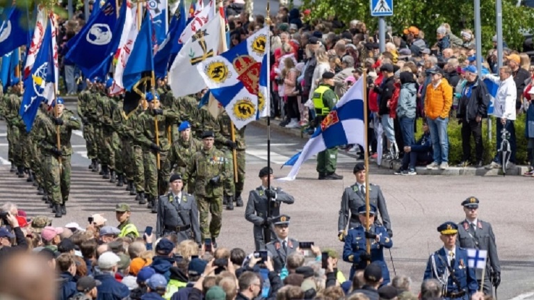Kuvassa paraatijoukko ja lippuja. Edessä Suomen valtiolippu. Ympärillä yleisöä.