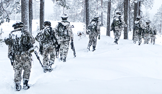 Talvimaisemassa kahdeksan talvivarustuksessa olevaa taistelijaa kävelemässä metsässä.
