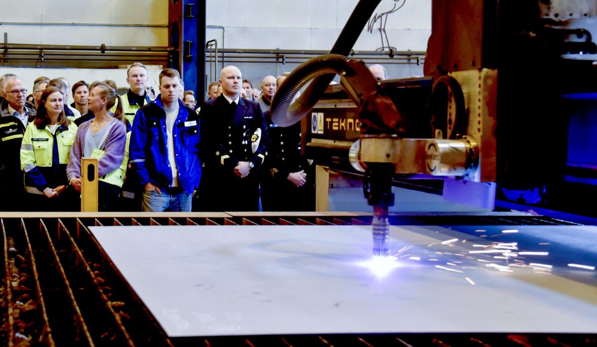 In the foreground, the ceremonial cutting of the steel plate, followed by a group of invited guests visible in the background.